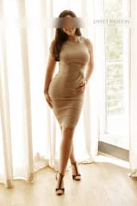 michelle high class escort köln vor einem fenster stehend in einem engen beigen kleid und schwarzen high heels, ihre dunkelbraunen langen haare sind offen und glatt